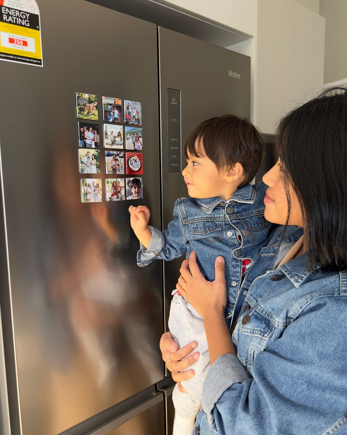 A mum holding her toddler at a fridge covered in a grid of square photo magnets
